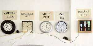 Verschiedene Wanduhren mit Zetteln darüber, wo drauf steht "retro Zeit", "Kommune Zeit" und anderes