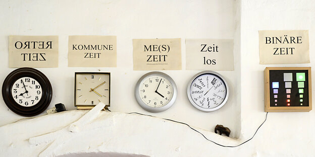 Verschiedene Wanduhren mit Zetteln darüber, wo drauf steht "retro Zeit", "Kommune Zeit" und anderes