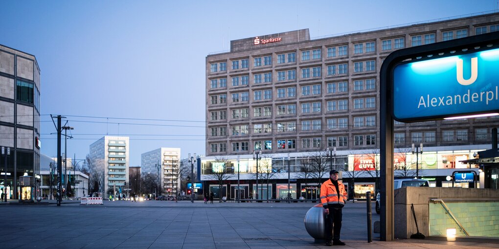 Der menschenleere Alexanderplatz in der Abenddämmerung: an einem Abfallbehälter steht ein Mann und raucht eine Zigarette