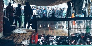 Das Bild wurde in einem Zelt aufgenommen. Im Vordergrund liegen leere Getränkedosen. Im Hintergrund stehen Männer in einer Schlange und warten.