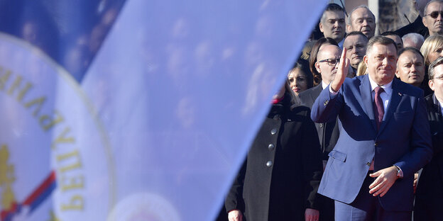 Milorad Dodik vor einer Menschenmenge, er winkt. Die Szene wird von einer bosnischen Fahne im Vordergrund bedeckt