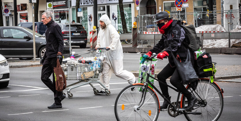 ann mit Schutzanzug und vermülltem Einkaufswagen überquert mit anderen eine Straße.