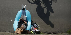 Eine Frau sitzt auf einem Luftkissen am Rheinufer und liest während ein Radfahrer vorbeifährt.