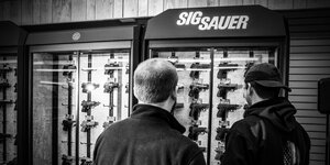 Ein schwarzweißbild: Menschen stehen vor einem Schrank mit Waffen von SIg Sauer