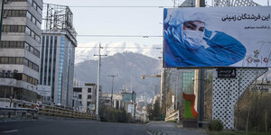 eine leere Straße, im Hintergrund Berge, am Rand ein Plakat mit einer Person mit Mundschutz