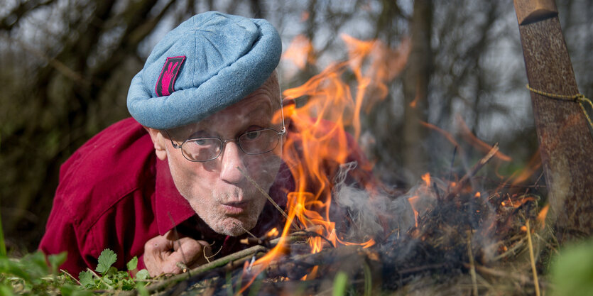 Eni Mann mit flacher runder Mütze bläst ein Feuer an