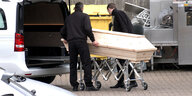 Zwei Männer in dunklen Anzügen schieben einen Sarg in einen Leichenwagen