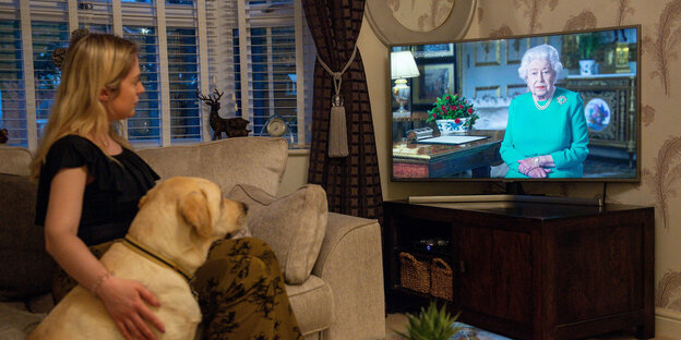 Eine Frau und ein Hund sitzen vor dem Fernseher. Auf dem Bildschirm ist die Queen zu sehen