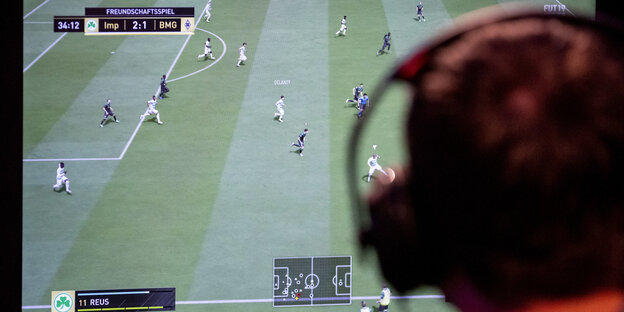 Ein Mensch mit Kopfhörer sitzt vor einem Bildschirm, auf dem ein virtuelles Fußballspiel gespielt wird.