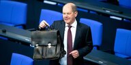Olaf Scholz hält seine Aktentasche und steht zwischen den Sitzen im Bundestag