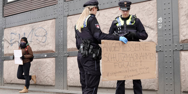 Vor einer Wand stehen zwei Polizisten mit einem Pappschlild von Demonstranten, links davon lehnt eine einzelne Demonstrantin an der Wand