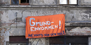 Ein orangefarbenes Transparent mit der Aufschrift "Grundeinkommen - Nahmt's von den Recihen" hängt an einer Fassade