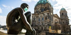 Ein Figur aus Bronze sitzt auf einer Ufermauer vor dem Berliner Dom und trägt Mundschutz.