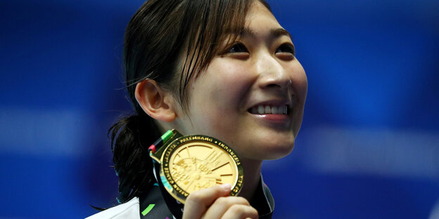Eine junge Frau hält eine Medaille und grinst.