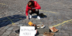 Frau malt mit Kreise auf den Boden. Neben ihr: abgestellte Schuhe und das Schild "Kapitalismus tötet"