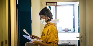 Eine Frau trägt eine Mundschutzmaske, hält Anschreiben in den Händen und steh im Flur des Gesundheitsamts Steglitz