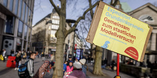 Im Vordergrund ein Demo-Pappschild, dass die Einführung eines Grundeinkommens fordert, im Hintergrund verschwommen Menschen in einer Fußgängerzone