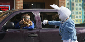 Ein als Osterhase verkleideter Mann winkt einem kleinen Jungen, der auf dem Schoß seines Vaters im Auto sitzt.
