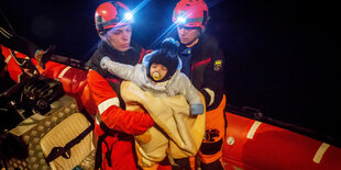 Zwei Männer in Schutzkleidung heben ein Baby von einem Boot