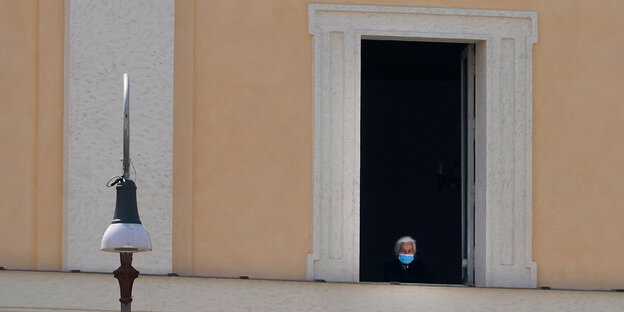 eine Frau mit Mundschutz schaut aus einem großen Fenster auf den Petersplatz