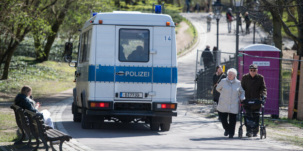 Ein Polizei-Mannschaftswagen fährt durch einen Park, rechts und links Spaziergänger und Menschen auf einer Parkbank Ein Polizei-Mannschaftswagen fährt durch einen Park, rechts und links Spaziergänger und Menschen auf einer Parkbank