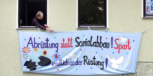 Ein Mann hängt ein Transparent aus seinem Wohnungsfenster. Darauf steht in bunter Schrift: "Abrüstung statt Sozialabbau! Spart endlich an der Rüstung!" Auf dem Transparent sind außerdem Blumen, Friedenstauben und eine zerbrochene Rakete aufgemalt.