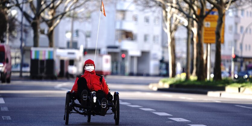 Ein Mann fährt auf einem Liegefahrrad über eine leere Straße, er trägt Mundschutz.