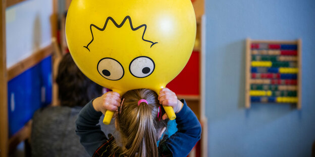 Ein kleines Mädchen von hinten, sie hält einen gelben Gummiball über ihren Kopf, dessen aufgemaltes, lächelndes Gesicht zeigt nach unten