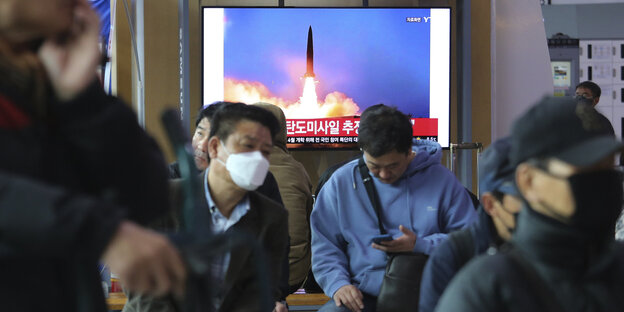 Menschen mit Mundschutz schauen auf ihre Handys, im Hintergrund steigt auf einem Fernseher eine Rakete in die Luft.