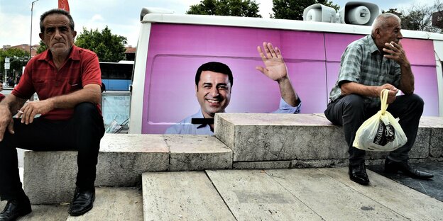 Zwei Männer sitzen auf Steinstufen, in der Mitte ein Plakat mit dem winkenden Politiker Selahattin Demirtas