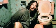 Ein Mann mit Bademantel und nassen Haaren sitzt neben einer Toilette.
