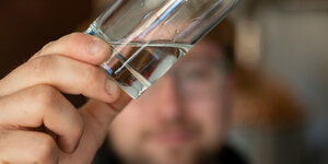 Ein Mann hält ein Schnapsglas mit einer transparenten Flüssigkeit vor die Kamera