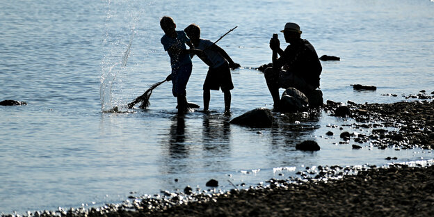 Zwei Kinder spielen am Ufer eines Sees, ein Erwachsener sitzt auf einem Stein und schaut zu