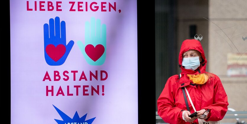 eine Frau mit Mundschutz steht neben einem Plakat