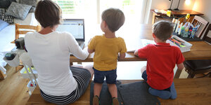 Von hinten ist eine Mutter zu sehen, die am Laptop arbeitet, neben ihr zwei Kinder