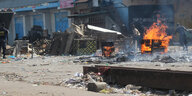 Menschen mit Mundschutz stehen hinter einer zerstörten und Marktständen, rechts im Bild lodert ein Feuer.