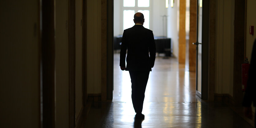 Olaf Scholz von hinten fotografiert, steht auf einem unbeleuchteten Flur.