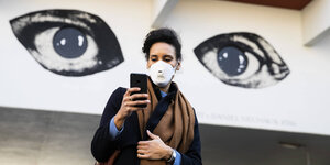 Eine Frau mit Mundschutz schaut auf ihr Smartphone, im Hintergrund sieht man große Augen an einer Wand.