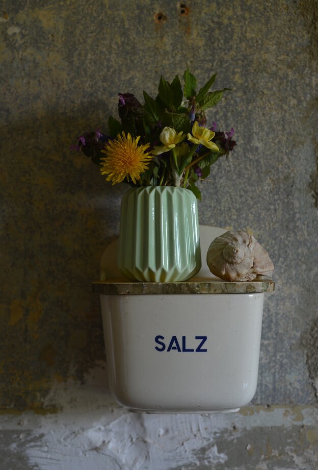 Ein altes Gefäß für Salz hängt an der Wand, auf diesem steht eine Vase mit Blumen