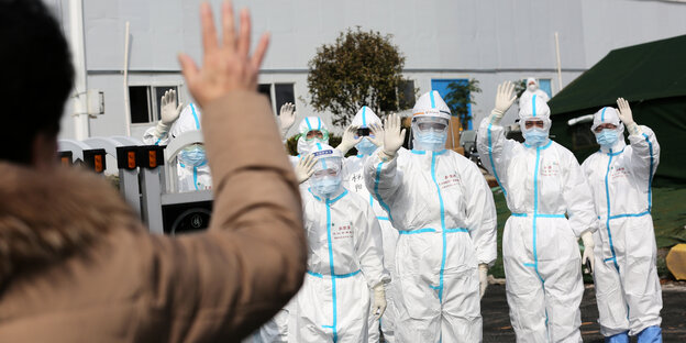 Menschen in Schutzanzügen winken einer Person in einer braunen Jacke. Menschen in Schutzanzügen winken einer Person in einer braunen Jacke.