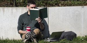 Ein junger Mann liest an eine Mauer gelehnt ein Buch.