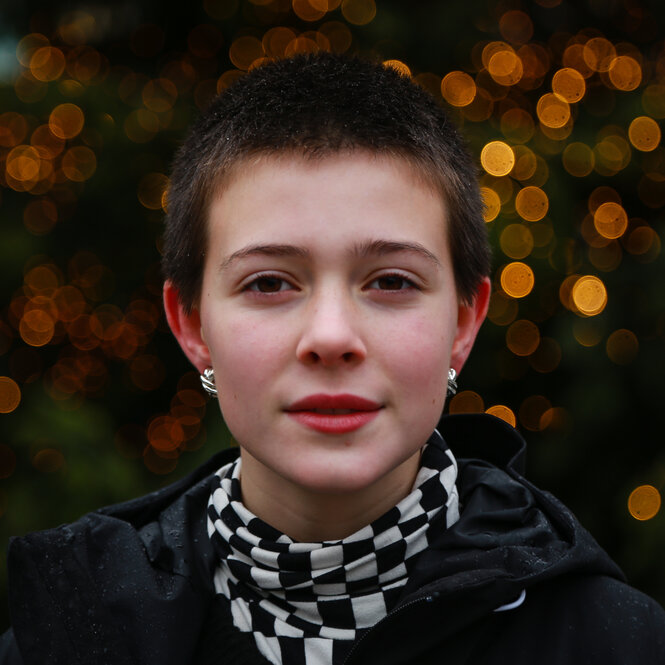 Maira Kellers, eine junge Frau mit kurzen dunklen Haaren. Sie trägt einen Schal und Ohrringe. Im Hintergrund ist ein Gebüsch oder Baum.