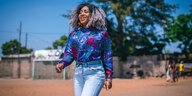 Die Rapperin Dama do Bling aus Mosambik geht über eine Straße. Sie trägt eine bunte, glänzende Bluse und eine Jeans. Ihre Haare trägt sie offen. Sie lacht.