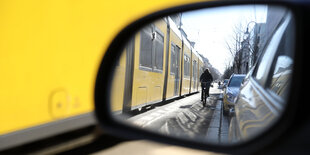 In einem Autorückspiegel sieht man einen Radfahrer und eine Straßenbahn.