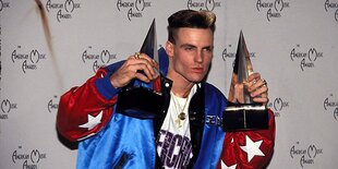 Ein Mann, der Rapper Vanilla Ice, in einer rot-blauen Ballonseide-Jacke posiert mit zwei pyramidenförmigen Trophäen