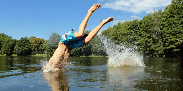 Eine Junge springt kopfüber in einen See mit Waldufer, der Kopf ist schon unter Wasser