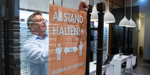 Ein Augenoptiker hängt in seinem Geschäft in der Mall of Berlin ein Schild mit der Aufschrift «Abstand halten! - Bleiben Sie gesund!» an die Eingangstür.