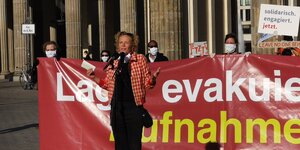 Schauspielerin Katja Riemann spricht auf einer kleinen Demo vor dem Brandenburger Tor, hinter ihr sind Menschen mit Atemmasken zu sehen, die das Banner "Lager evakuieren - Aufnahme jetzt" hochhalten