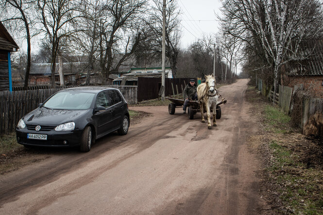 Eine staubige Straße in einem Dorf, an der Seite steht ein Auto, dem Betrachter kommt eine Kutsche mit einem Pferd entgegen.