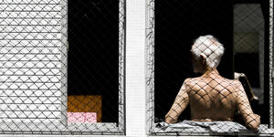 ein älterer Mann sonnt sich den Rücken an einem vergitterten Fenster sitzend